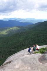 3 hikers on a large, smooth, rounded rock with green mountains in the background